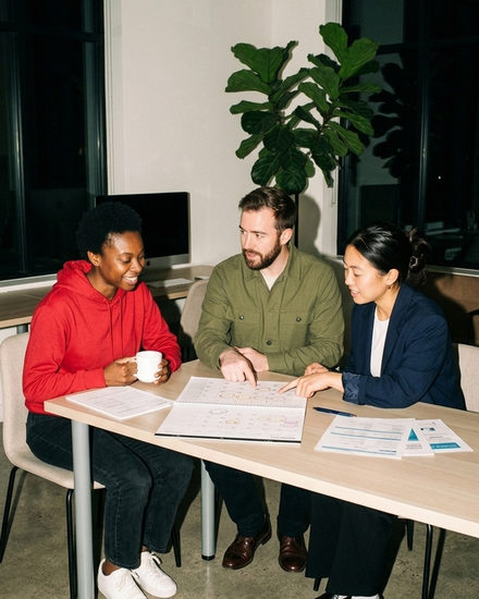 Three colleagues at a desk in an office, looking together at a wall planner open in front of them with payslips beside it.